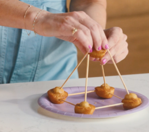 Four Little Bites Muffins spread out on a plate and connected by popsicle sticks, with popsicle sticks also coming out of all four tops and a pair of hands connecting them all at the top with a final Little Bites Muffin to create a pyramid. 